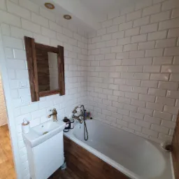 Biała łazienka z wanną obudowaną drewnem, umywalką na szafce i lustrem w drewnianej ramie, płytki typu metro na ścianach, złota armatura.