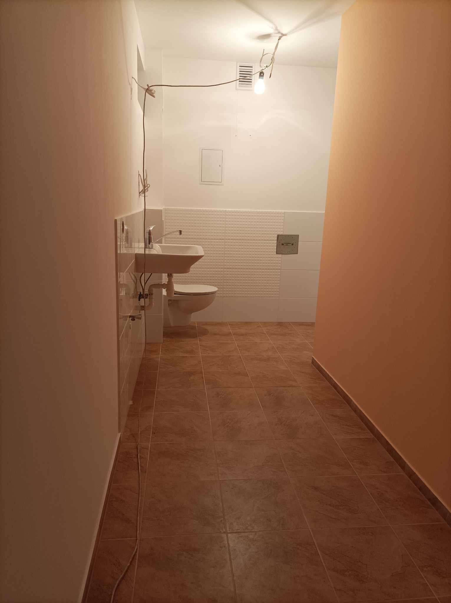 Wnętrze łazienki w trakcie remontu, widoczne brązowe płytki na podłodze, biała toaleta i umywalka, ściany w kolorze brzoskwiniowym i białym, surowa żarówka na kablu.