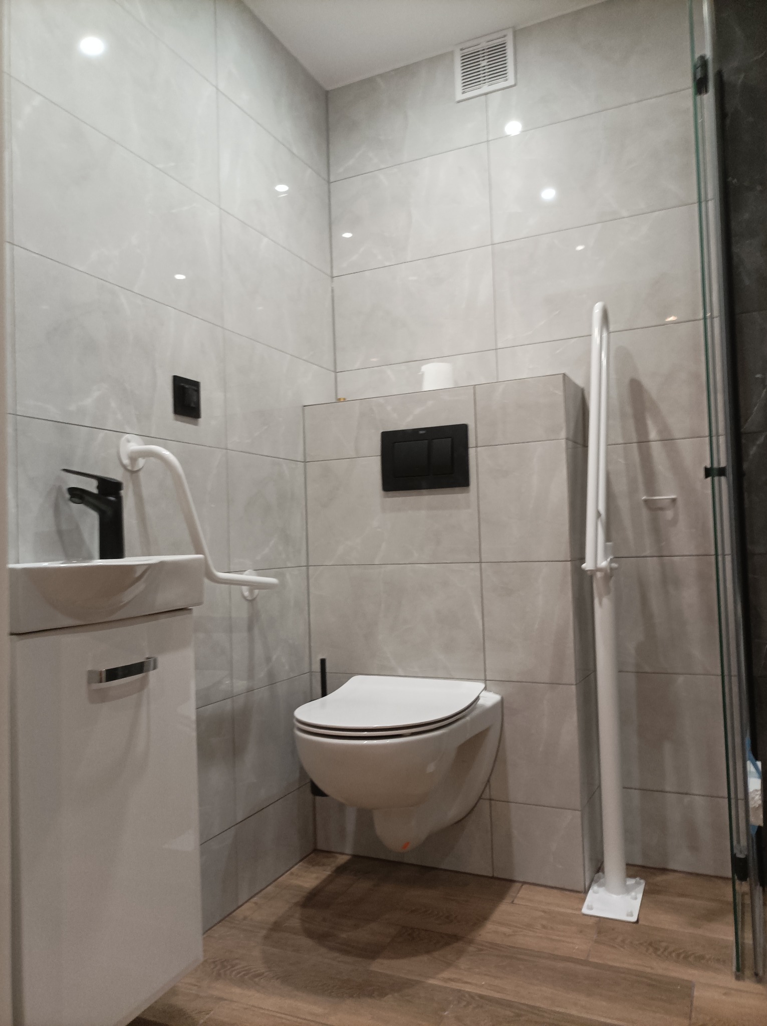 Nowoczesna łazienka z wiszącą toaletą, umywalką na szafce i uchwytami dla osób z niepełnosprawnościami, wykończona szarymi płytkami imitującymi marmur i drewnianą podłogą.