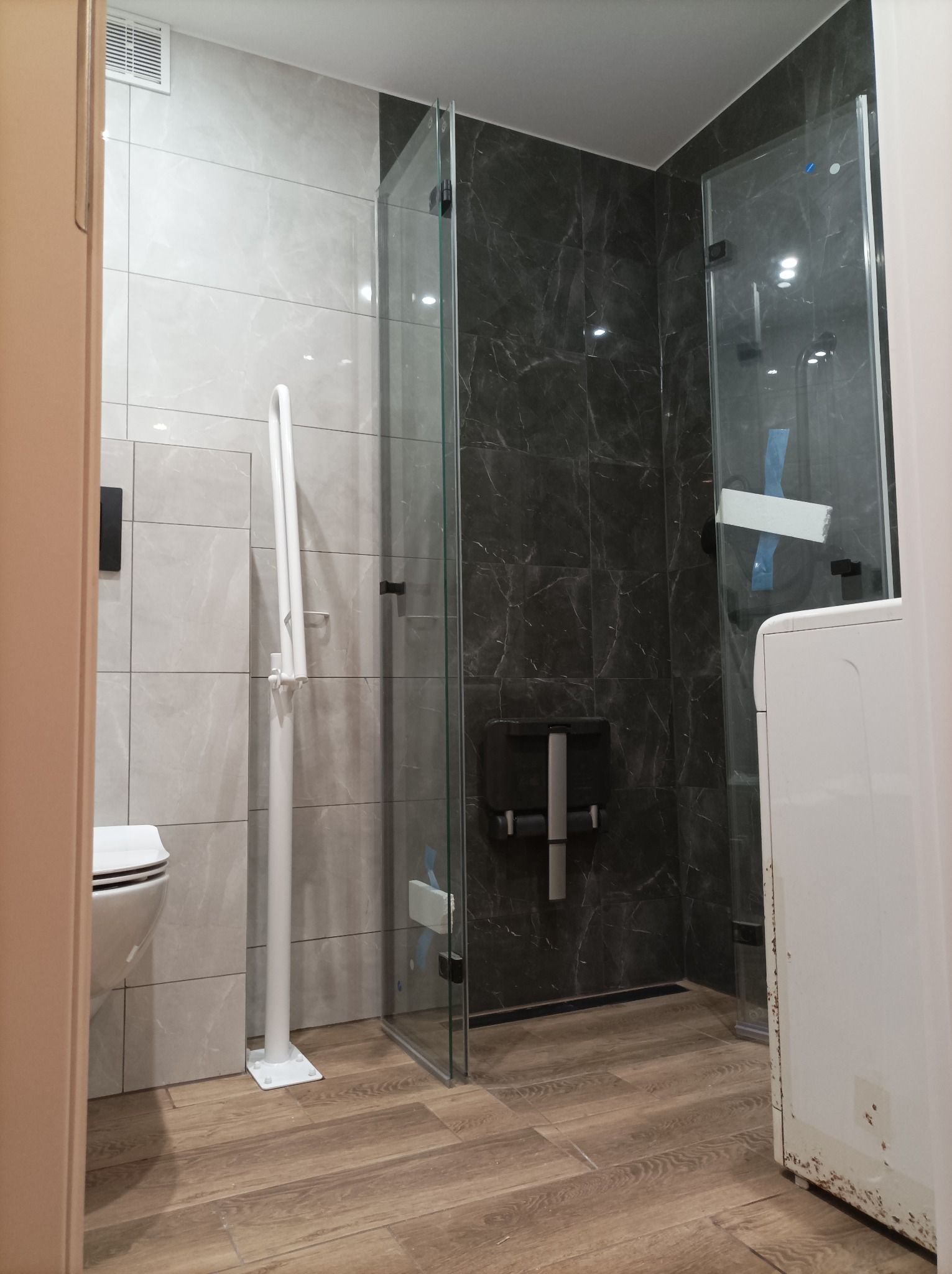 Wykończona łazienka z kabiną prysznicową typu walk-in, płytkami imitującymi marmur w odcieniach szarości, drewnopodobną podłogą, toaletą podwieszaną oraz uchwytem dla osób niepełnosprawnych...