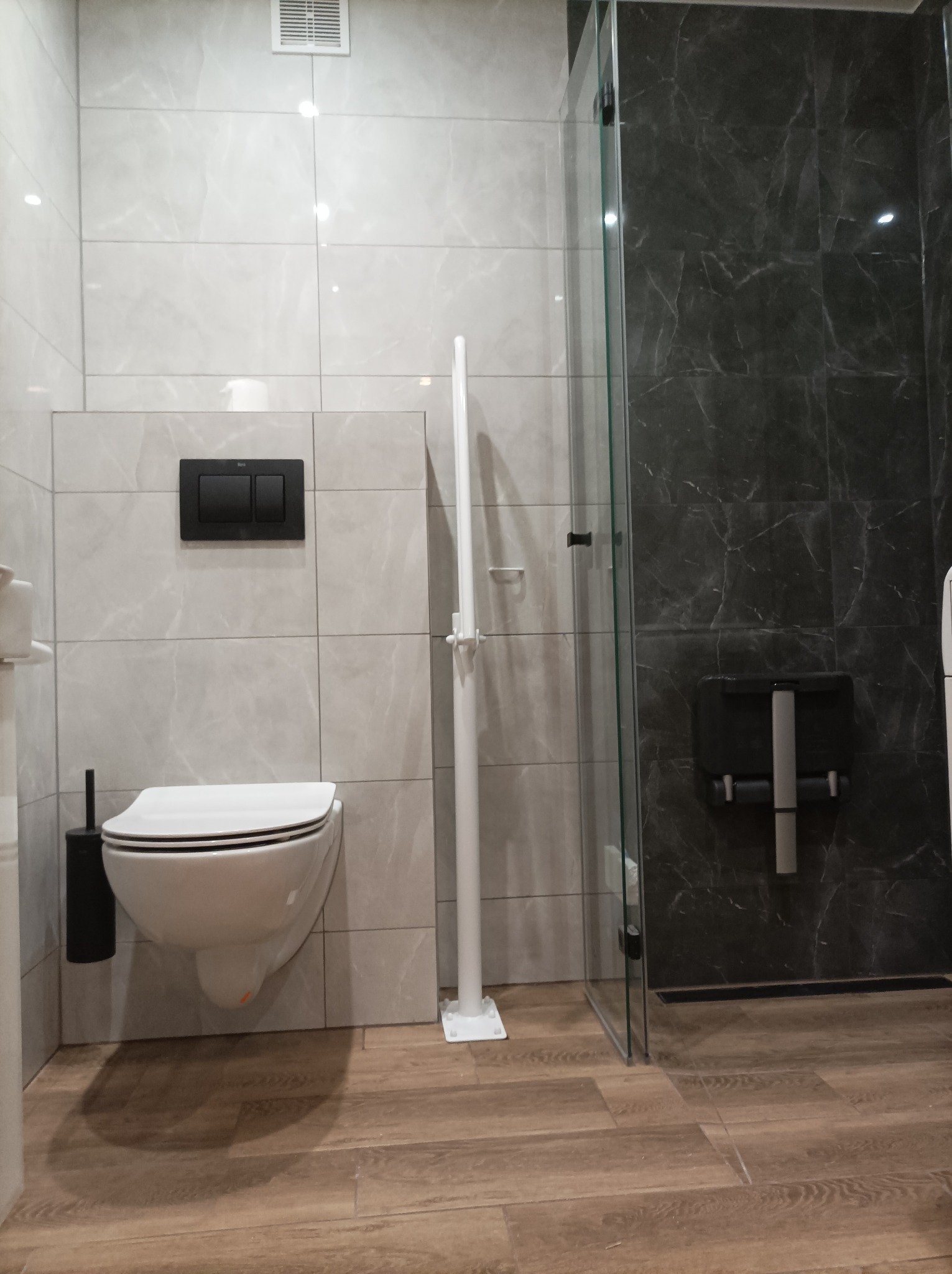 Nowoczesna łazienka z podwieszaną toaletą, czarnym przyciskiem spłukiwania, kabiną prysznicową z ciemnymi marmurowymi płytkami i drewnopodobną podłogą.