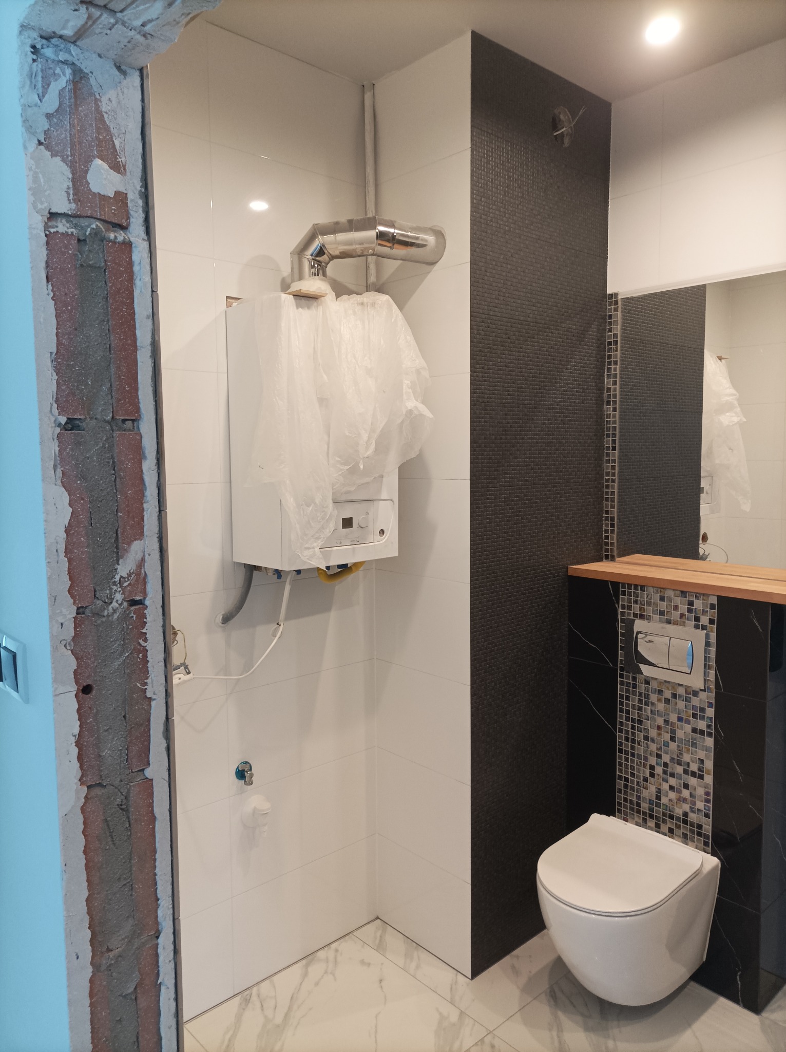 Wnętrze łazienki w trakcie remontu: widoczny wiszący sedes, fragment ściany z czarną mozaiką, białe płytki, piec gazowy osłonięty folią, surowy fragment ściany z cegły i niedokończone obramowanie...