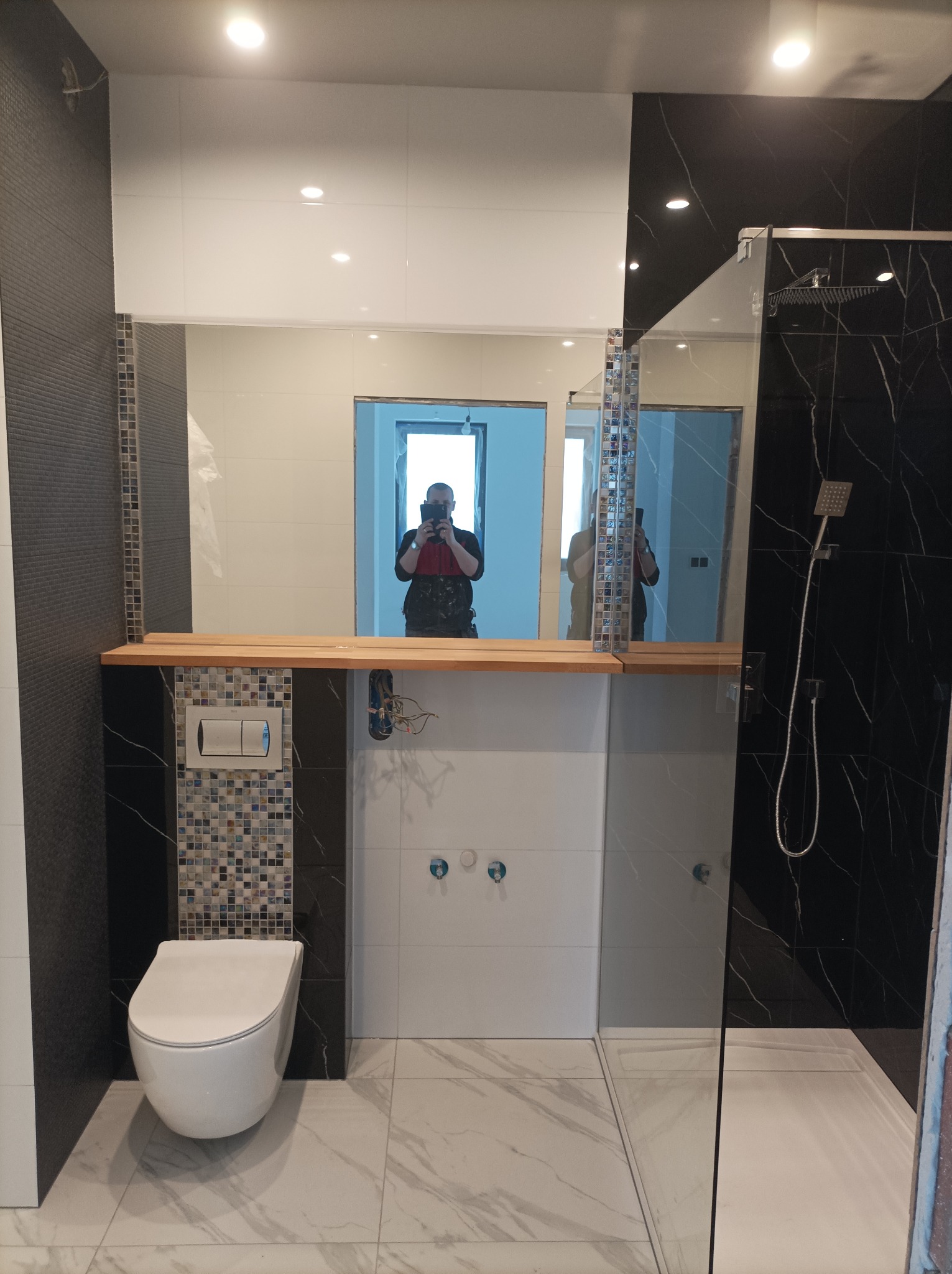 Nowoczesna łazienka w trakcie wykańczania, z wiszącą toaletą, czarną i białą glazurą, drewnianą półką pod lustrem i widocznym odbiciem fotografa.