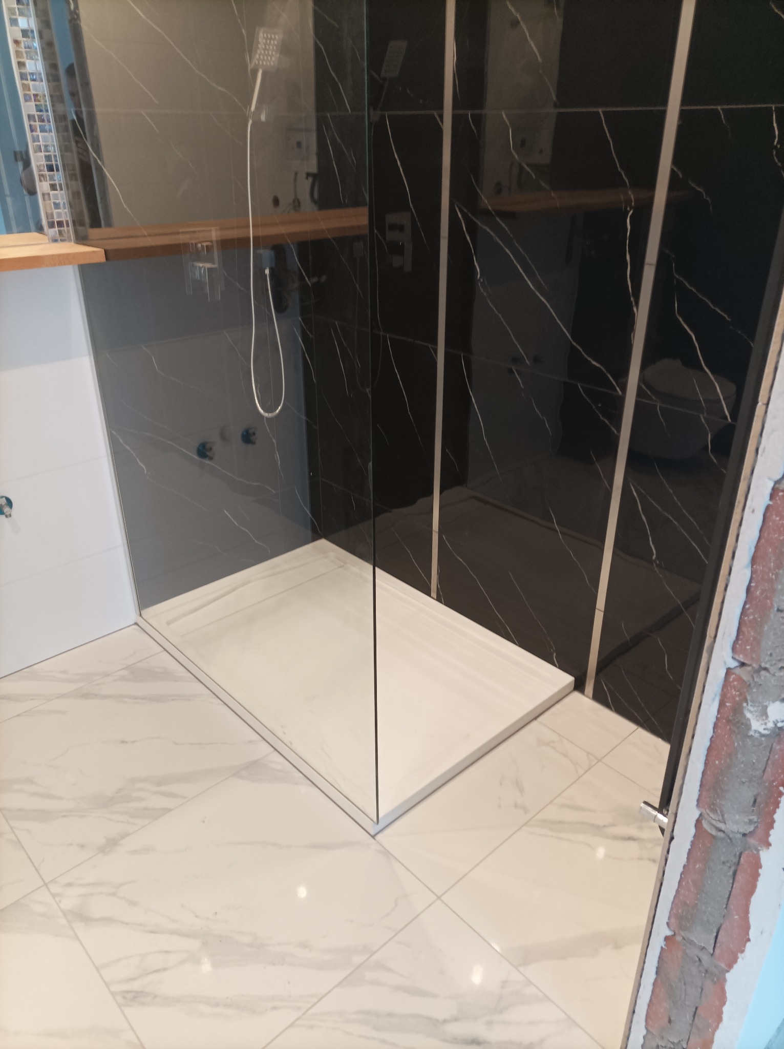 Nowoczesna kabina prysznicowa walk-in z czarnymi płytkami imitującymi marmur i białym brodzikiem, widok z góry na wykończoną łazienkę z marmurową podłogą i drewnianą półką.