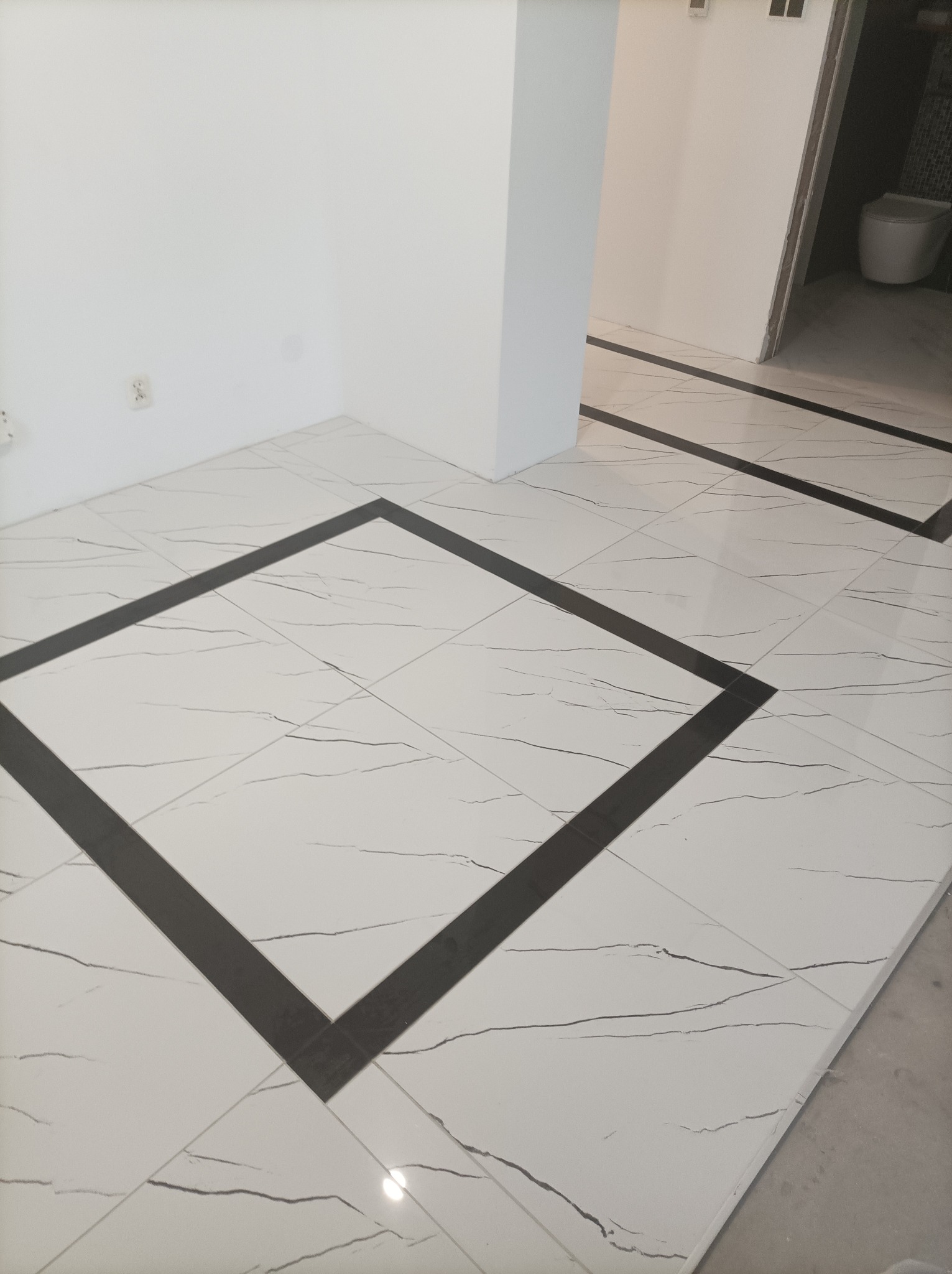 Wykończona podłoga z białych płytek imitujących marmur z czarnymi, kwadratowymi obramowaniami, widok na łazienkę z toaletą.
