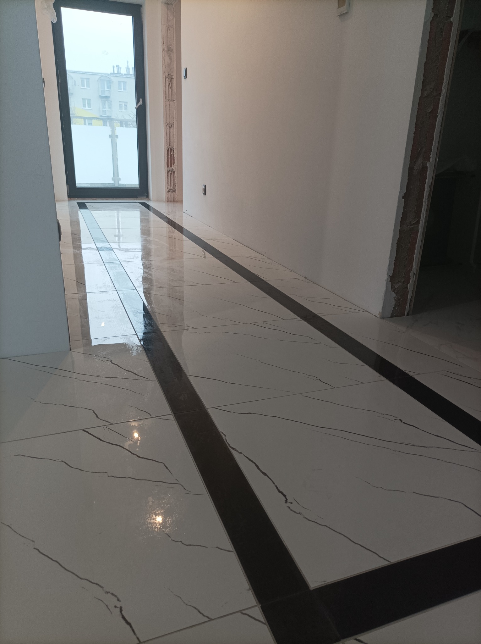 Błyszcząca, biała podłoga z marmurowym wzorem i czarnymi pasami dekoracyjnymi, świeżo ułożona w pustym pomieszczeniu z oknem wychodzącym na budynek mieszkalny.