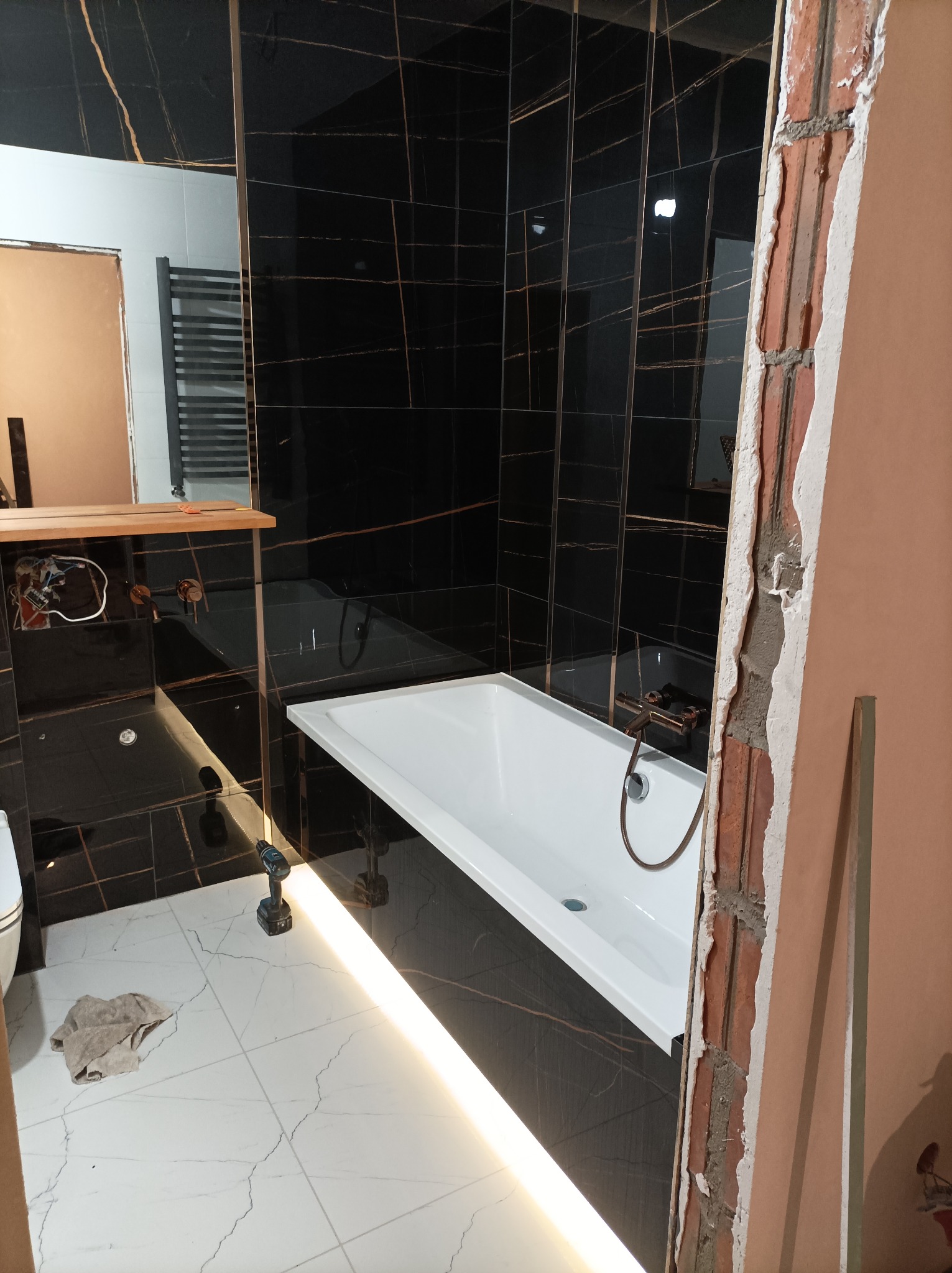 Wnętrze łazienki w trakcie prac wykończeniowych: biała wanna, czarne płytki z złotymi żyłami na ścianach, biała marmurowa podłoga, widoczna surowa ściana z cegły, wiertarka na podłodze i szmata obok