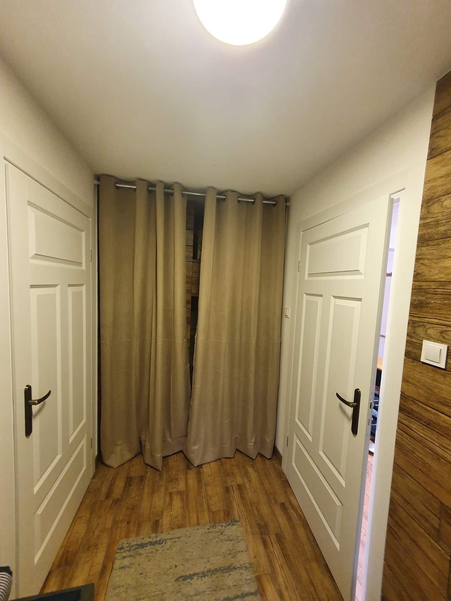Wnętrze korytarza z dwiema białymi, klasycznymi drzwiami po bokach i zasłoniętym wejściem na wprost beżowymi zasłonami na metalowym karniszu, drewniana podłoga i fragment dywanu w dolnej części kadru.