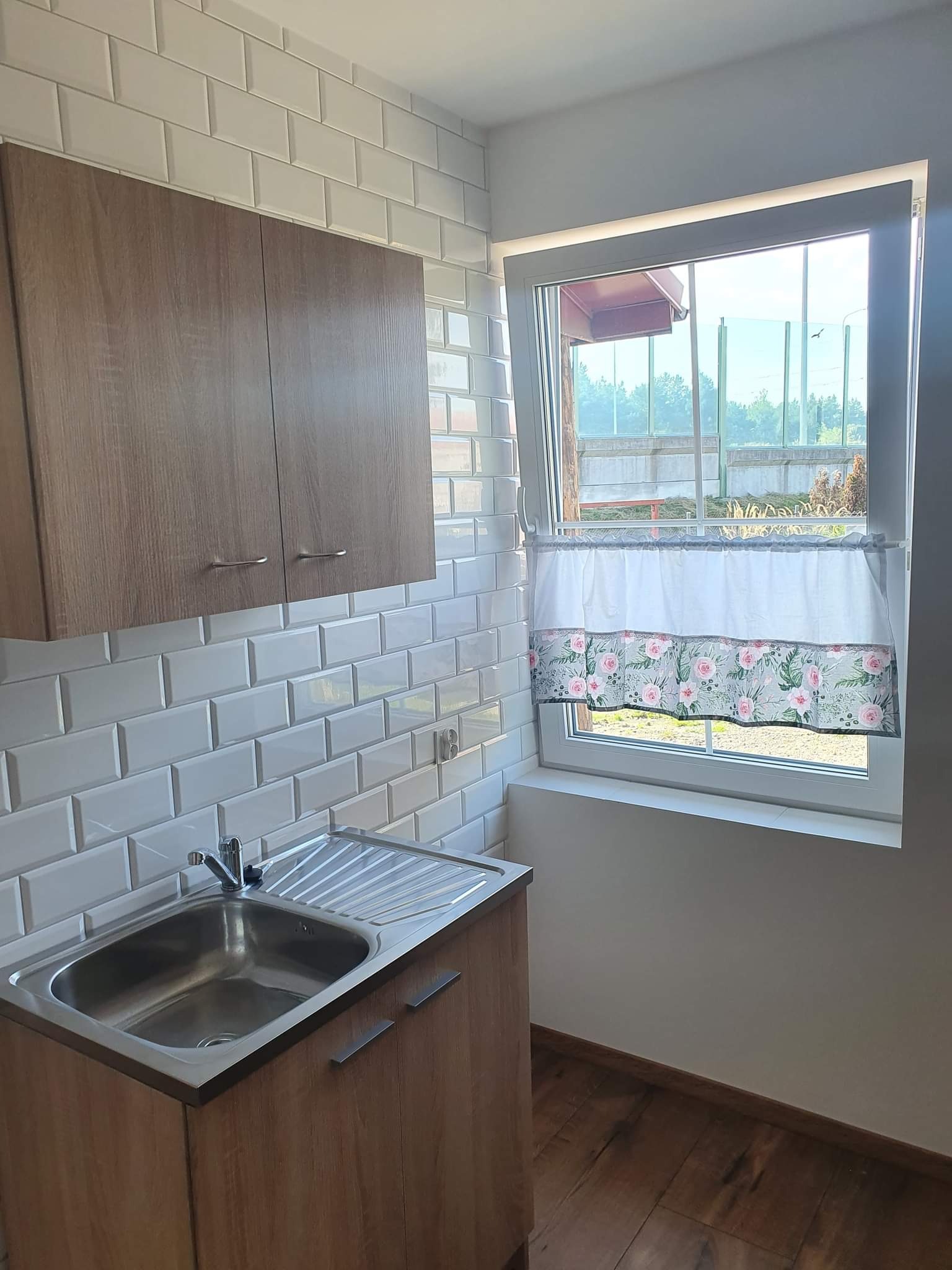 Umywalka w zabudowie z drewnopodobnymi szafkami, białe płytki imitujące cegłę na ścianie, okno z krótką zasłonką w kwiaty, widok na zieleń za oknem, drewniana podłoga.