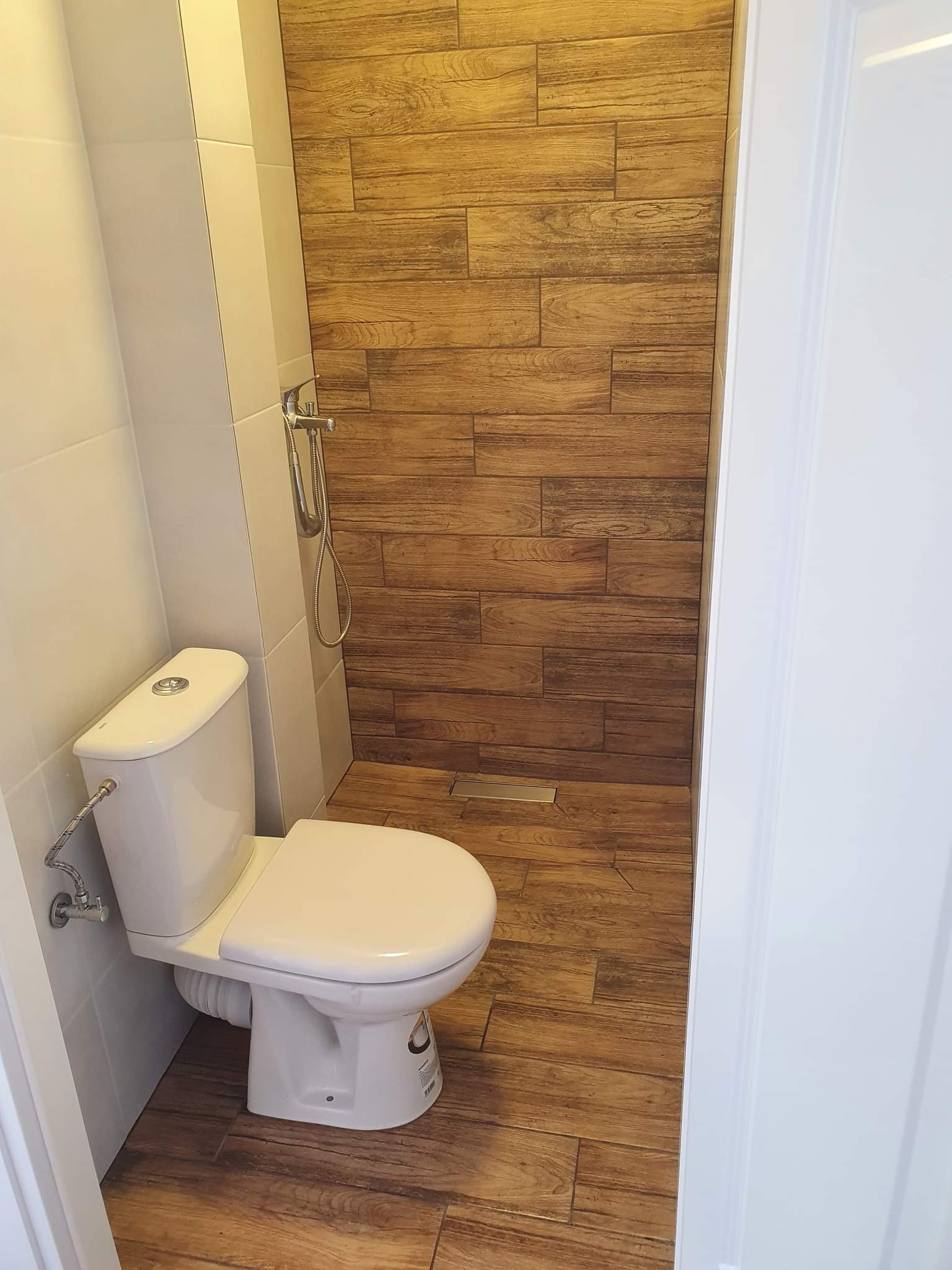 Nowoczesna toaleta z drewnopodobnymi płytkami na ścianie prysznicowej i podłodze, biała armatura, chromowana słuchawka prysznicowa.