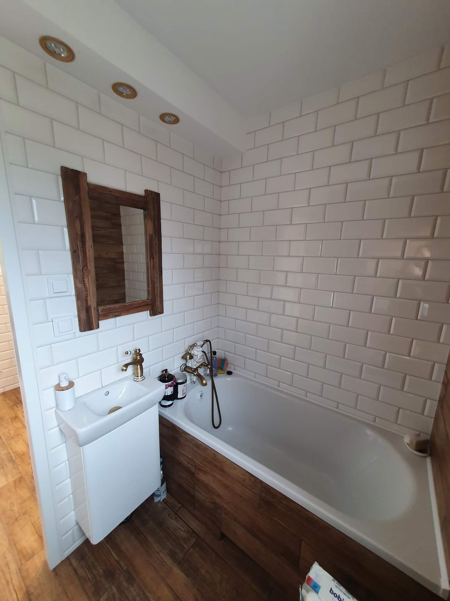 Biała łazienka z wanną obudowaną drewnem, umywalką na szafce i lustrem w drewnianej ramie, płytki typu metro na ścianach, złota armatura.