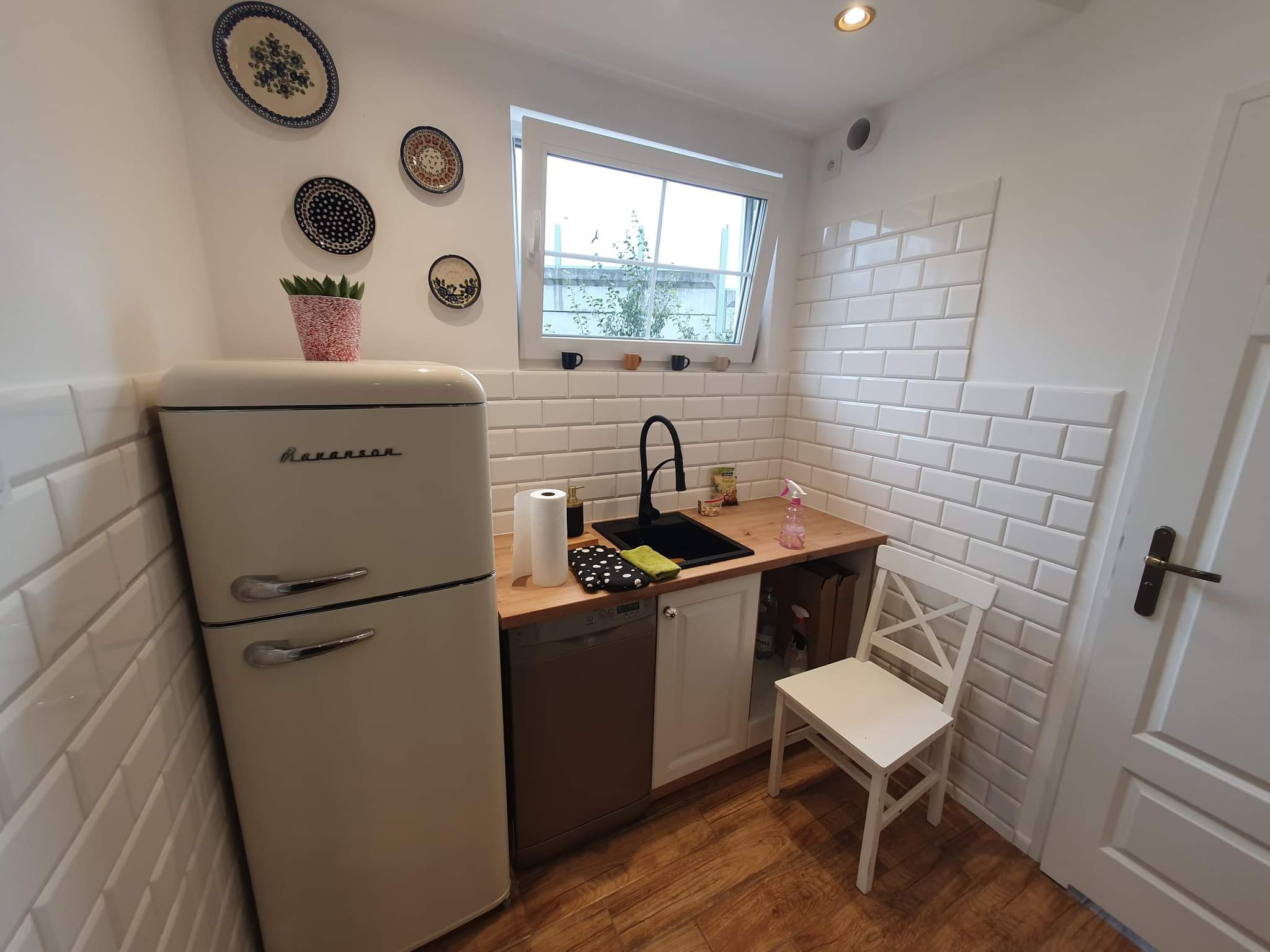 Kuchnia w stylu retro z beżową lodówką, drewnianym blatem, czarnym zlewem i białymi płytkami imitującymi cegłę, ozdobiona dekoracyjnymi talerzami na ścianie.