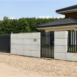 Nowoczesne ogrodzenie z przesuwną bramą w kolorze antracytowym, wykonane z betonowych bloczków i pionowych metalowych sztachet, z widocznym logo firmy na elemencie ogrodzenia.
