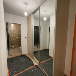 Montaż przesuwnej szafy z lustrzanymi drzwiami w trakcie prac wykończeniowych, podłoga zabezpieczona folią ochronną, widoczne niedociągnięcia w wykończeniu ścian i sufitu, żarówka bez klosza.