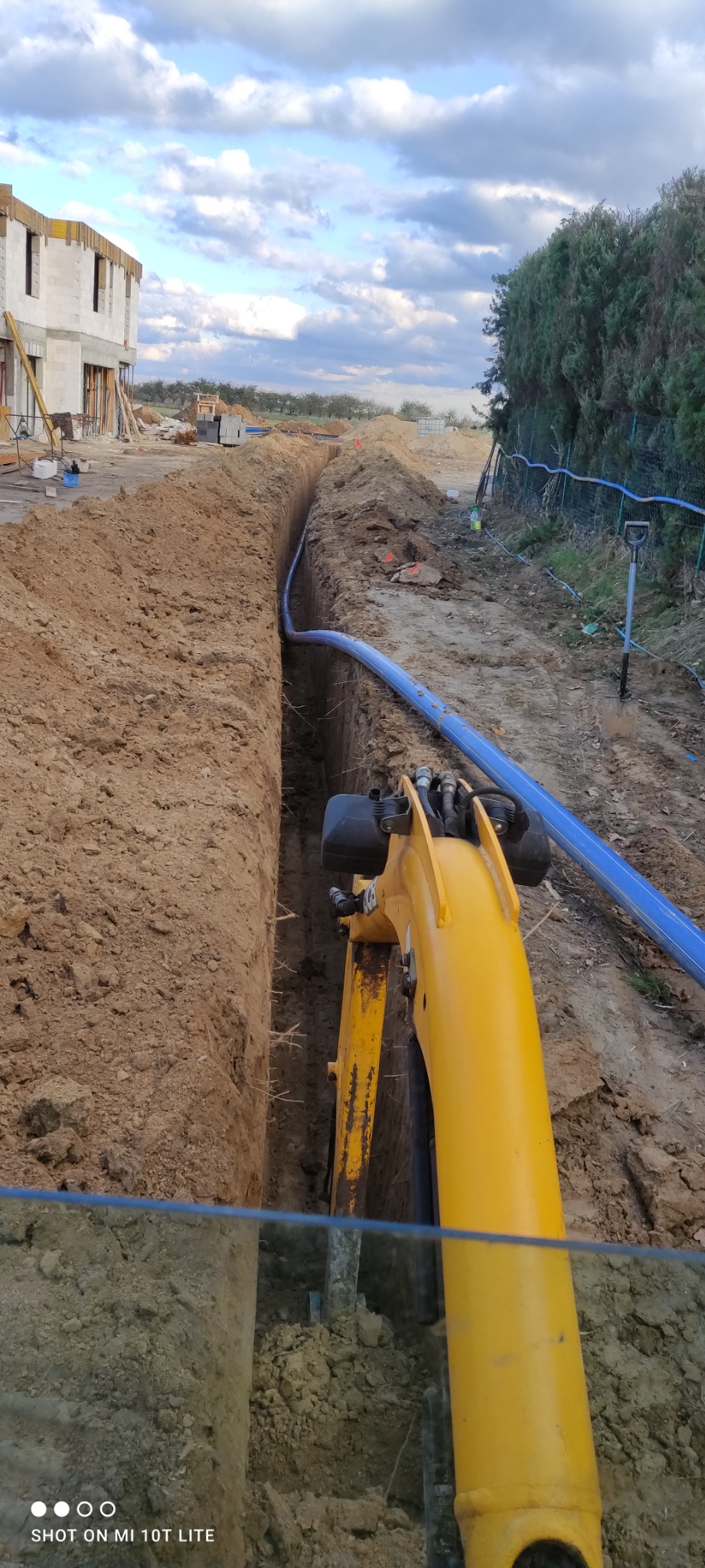 Żółta koparka umieszcza niebieski rurociąg w wykopanym wąskim rowie na placu budowy, w tle widać budynek w stanie surowym i drzewa.