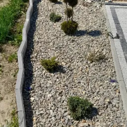 Wykończony fragment ogrodu z kamienną rabatą, ozdobnymi krzewami i obrzeżami z kostki, widok z góry na świeżo zaaranżowaną przestrzeń.