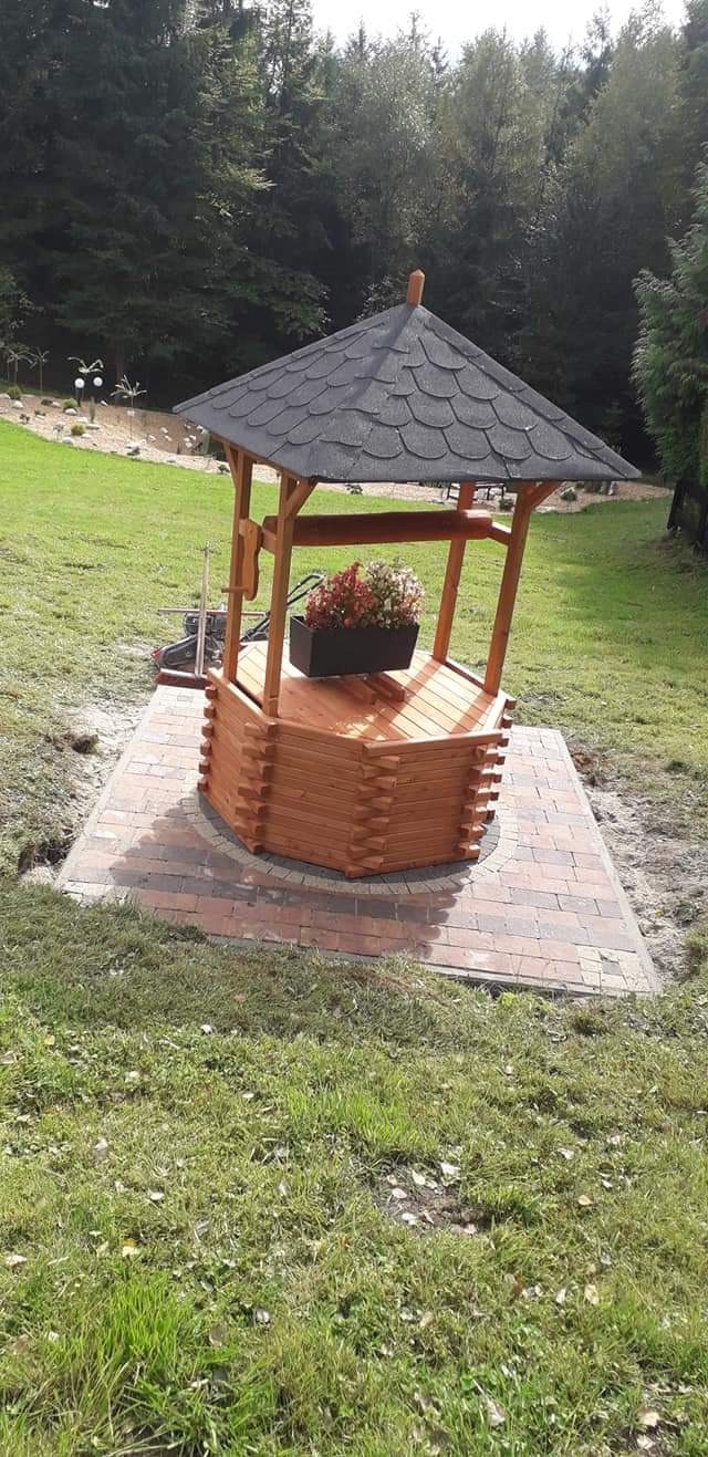 Drewniana studnia ogrodowa z dachem pokrytym gontem, umieszczona na kostce brukowej, z ozdobną skrzynką kwiatową wewnątrz, na tle zielonego trawnika i lasu.