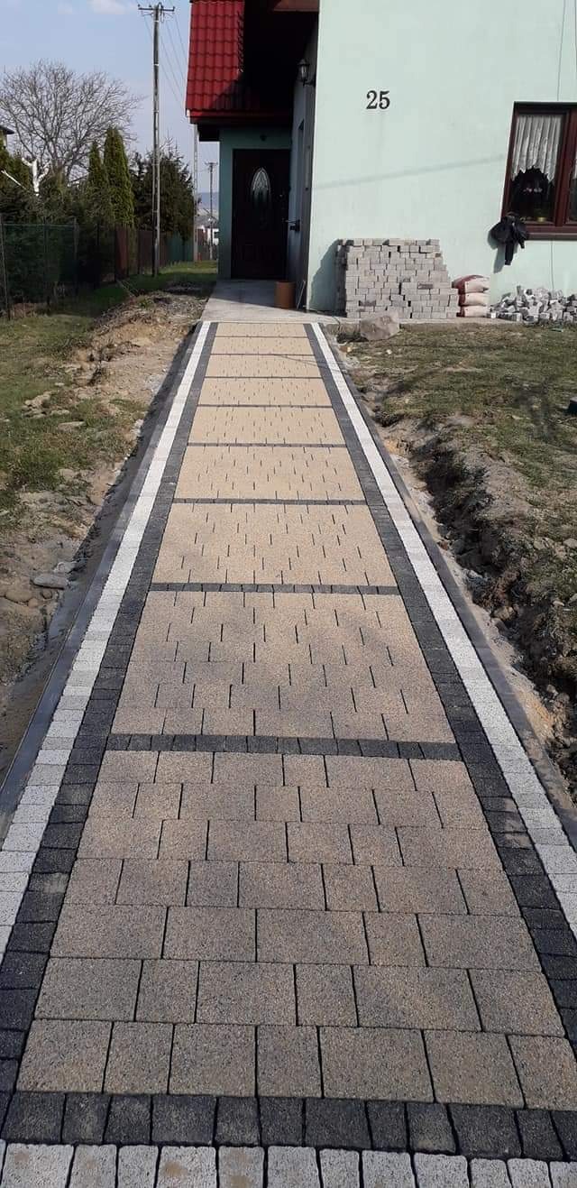 Ułożony chodnik z kostki brukowej w odcieniach beżu i szarości prowadzący do domu z numerem 25, widoczne obrzeża w kolorze białym i ciemnoszarym, w tle stos kostki brukowej i worki z materiałem.