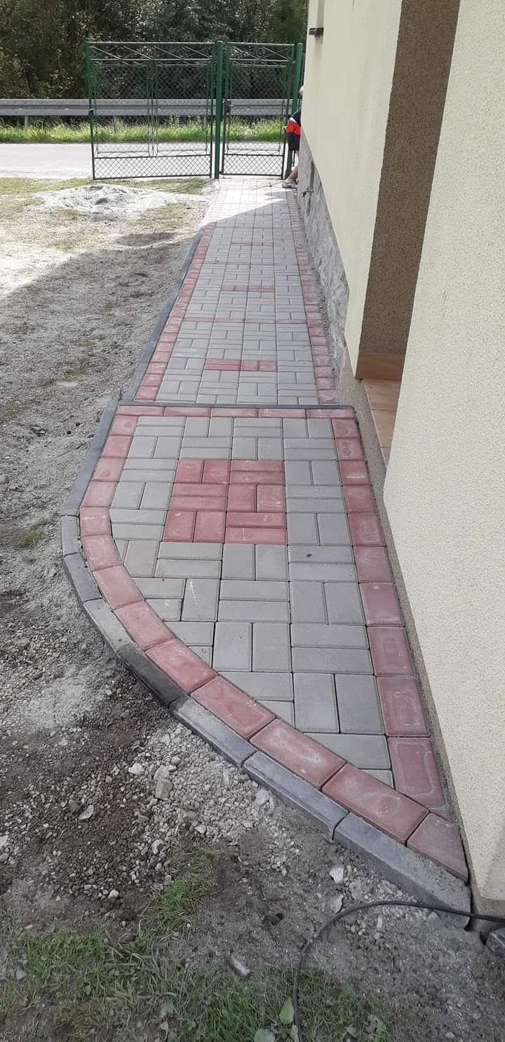 Ułożona ścieżka z szarej i czerwonej kostki brukowej przy ścianie budynku. Krawężnik z ciemnoszarej kostki. Widoczny fragment trawnika i ogrodzenia w tle.