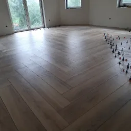 Ułożona pod kątem jasnobrązowa podłoga z płytek imitujących drewno, z widocznymi niebieskimi i czerwonymi elementami poziomującymi, w jasnym pomieszczeniu z oknami.