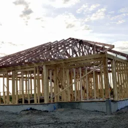 Surowy szkielet domu z drewna na fundamencie betonowym, widok na otwarte pola i zachmurzone niebo.