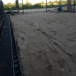 Przygotowany teren pod trawnik z widocznymi śladami po pracy, czarną geowłókniną na brzegu i pracownikiem w tle.