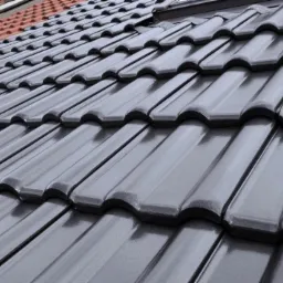 Błyszcząca, grafitowa dachówka ceramiczna ułożona rzędami, z fragmentem czerwonej dachówki w tle, widziana pod kątem.