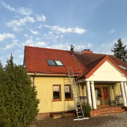 Pracownik w uprzęży na drabince dachowej czyści czerwoną dachówkę domu jednorodzinnego z poziomu dachu, z widocznym strumieniem wody i parą, na tle błękitnego nieba z chmurami.