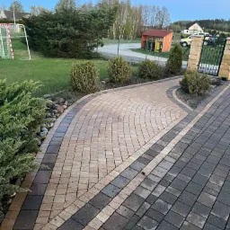 Ułożona z kostki brukowej ścieżka w ogrodzie, z widocznymi różnymi odcieniami kostki, obsadzona krzewami i kamieniami, prowadząca do bramy wjazdowej.