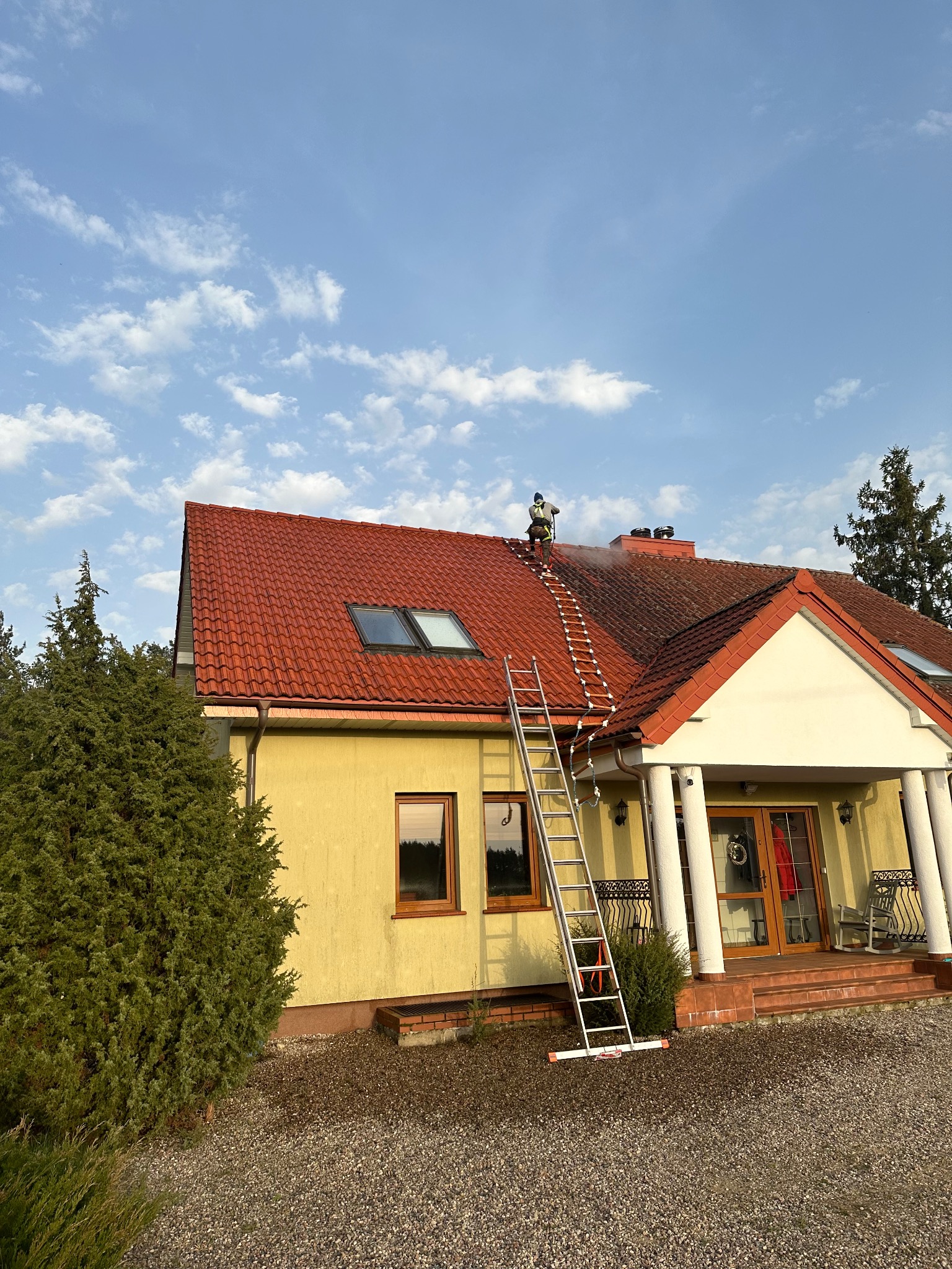 Pracownik w uprzęży na drabince dachowej czyści czerwoną dachówkę domu jednorodzinnego z poziomu dachu, z widocznym strumieniem wody i parą, na tle błękitnego nieba z chmurami.