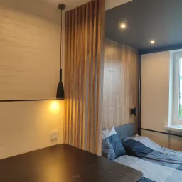 Nowoczesna sypialnia z drewnianą ścianką działową z pionowych listew, wisząca czarna lampa, drewniane panele ścienne i ł&oacute;żko z szaro-niebieską pościelą.