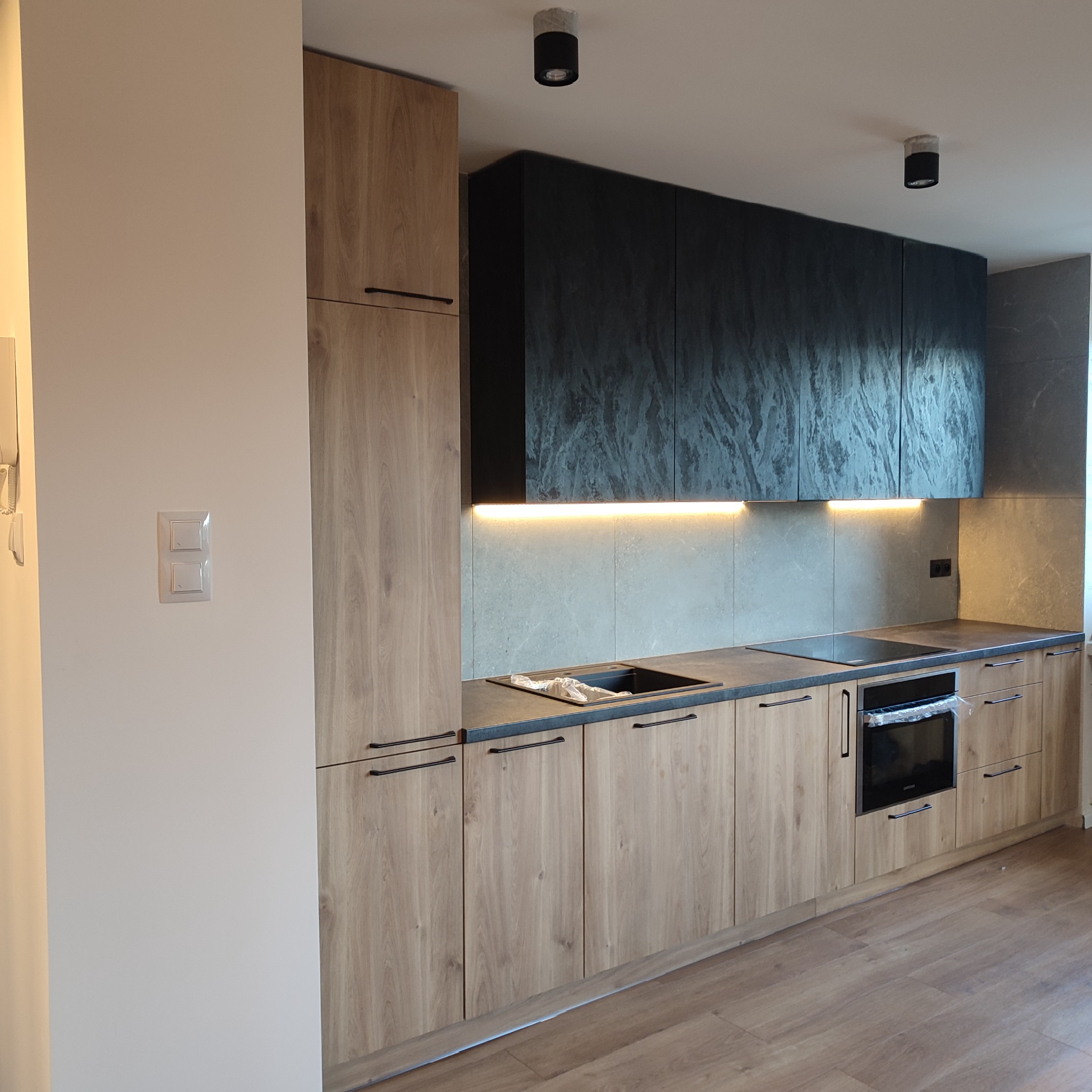 Nowoczesna kuchnia z frontami w odcieniach drewna i grafitowymi szafkami wiszącymi, podświetlona listwą LED, z czarnym zlewem i płytą indukcyjną, jeszcze w folii ochronnej.