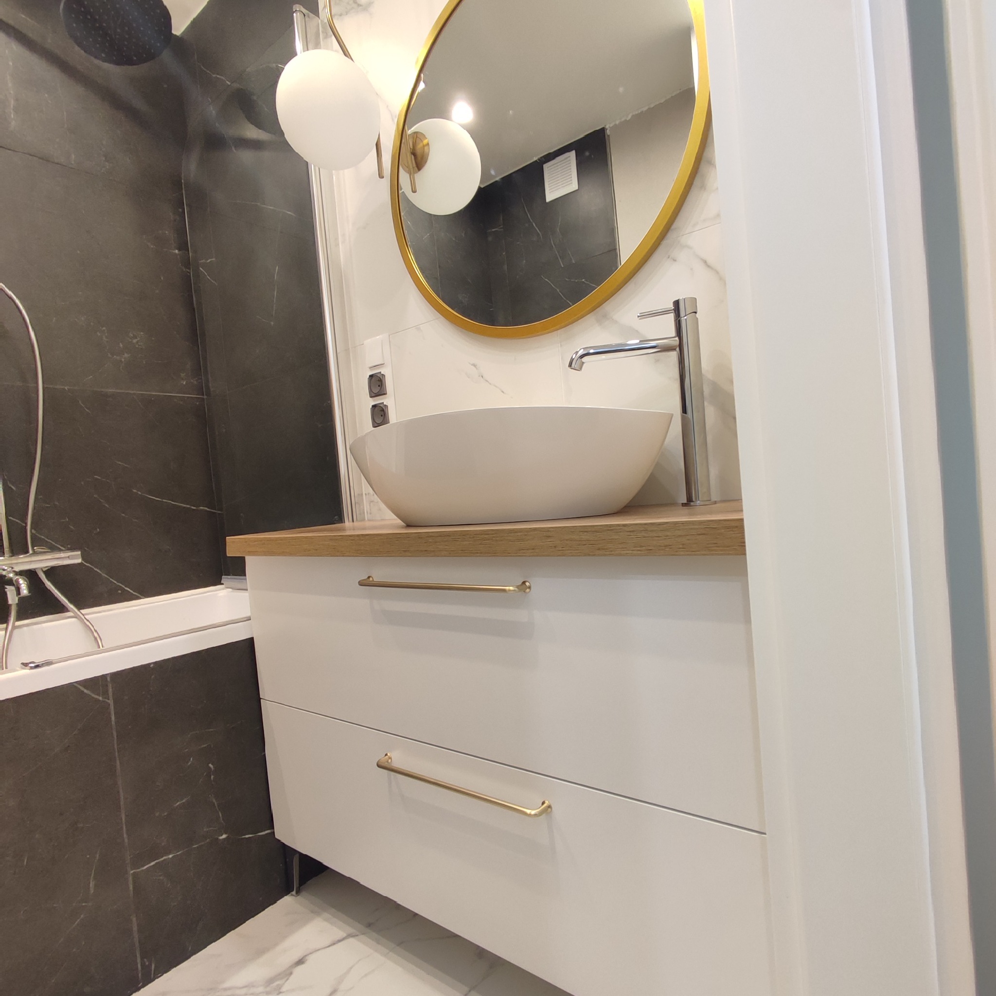 Nowoczesna łazienka z owalną umywalką nablatową, baterią chromowaną, okrągłym lustrem w złotej ramie, szafką z szufladami i złotymi uchwytami, oraz czarno-białymi płytkami imitującymi marmur.