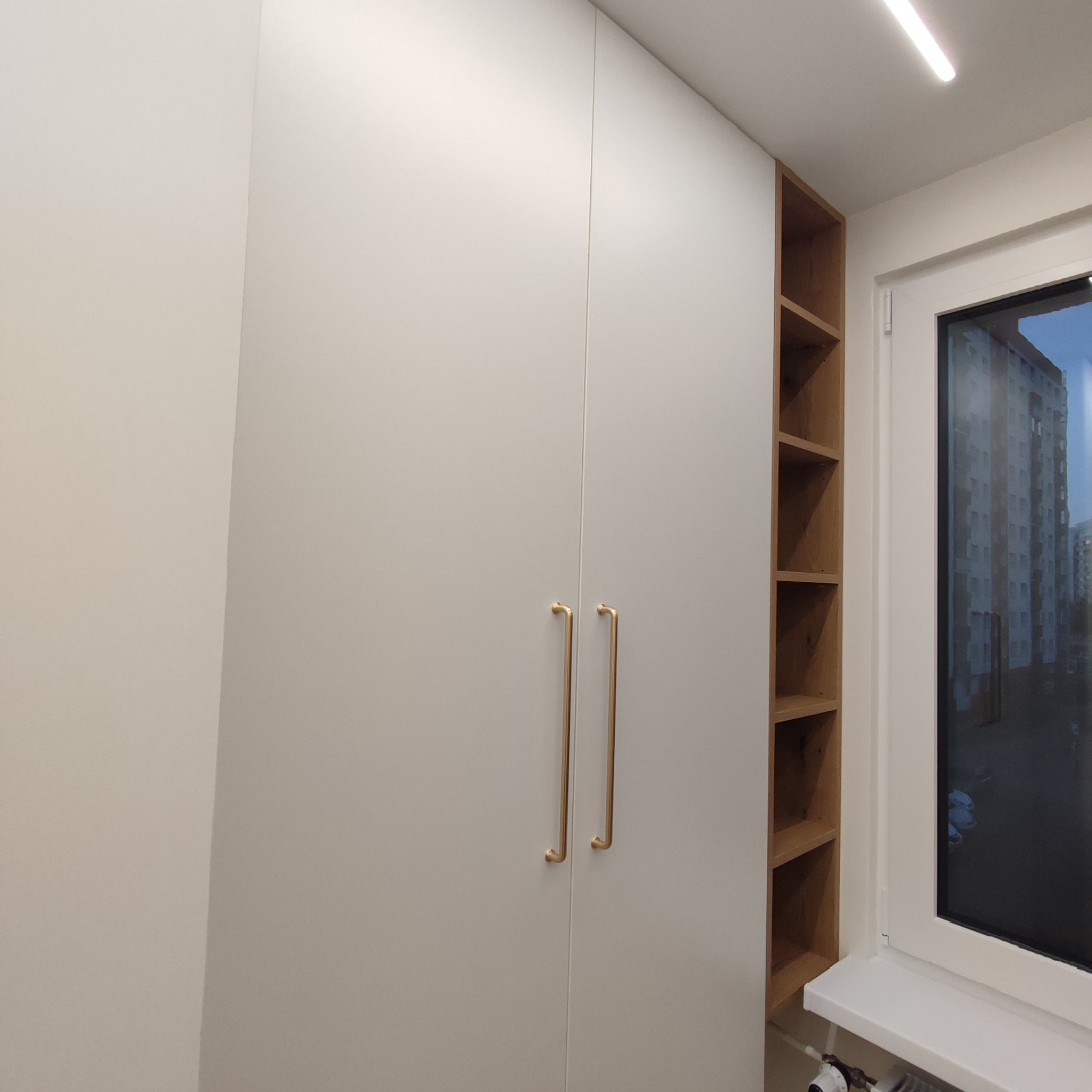 Biała szafa wnękowa z złotymi uchwytami, przylegająca do drewnianej półki wnękowej przy oknie z widokiem na blok mieszkalny.