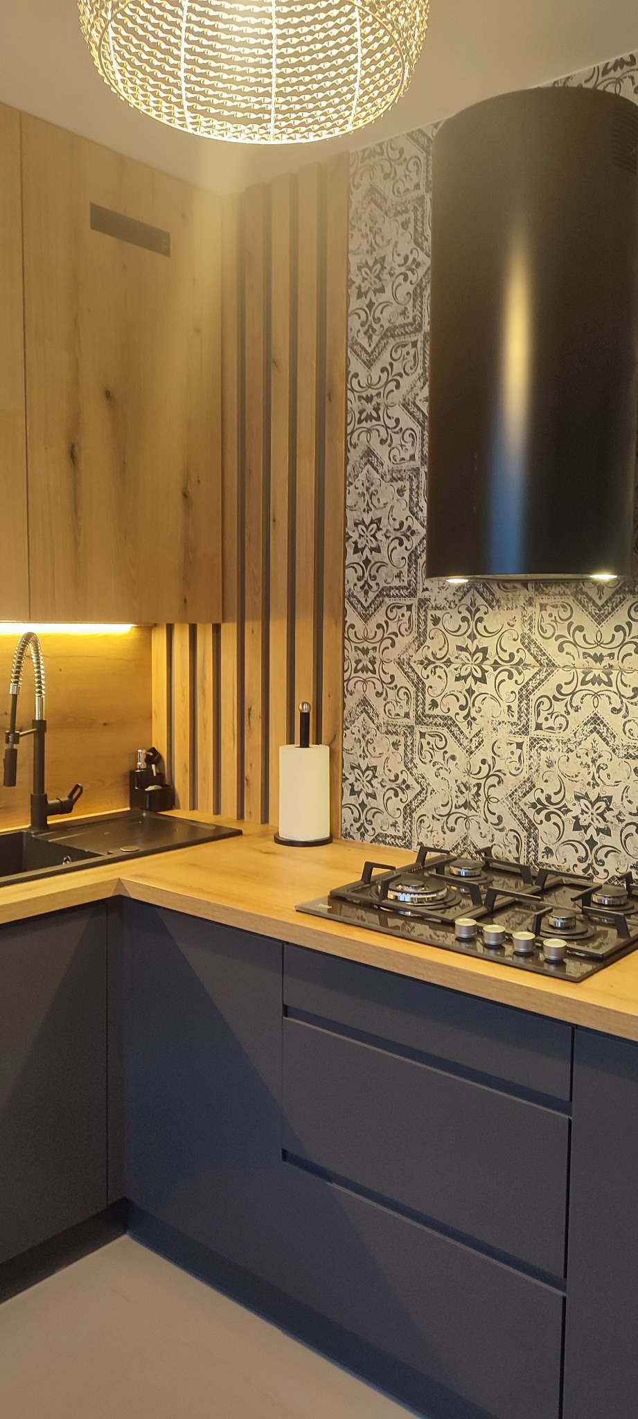 Fragment nowoczesnej kuchni z ciemnymi szafkami z płaskimi frontami, drewnianym blatem, czarną armaturą i płytą gazową; na ścianie dekoracyjne płytki w stylu patchwork oraz drewniane lamele.