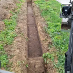 Wąski wykop liniowy w ziemi, widok z kabiny koparki, w tle zaparkowane ciężarówki i trawnik.