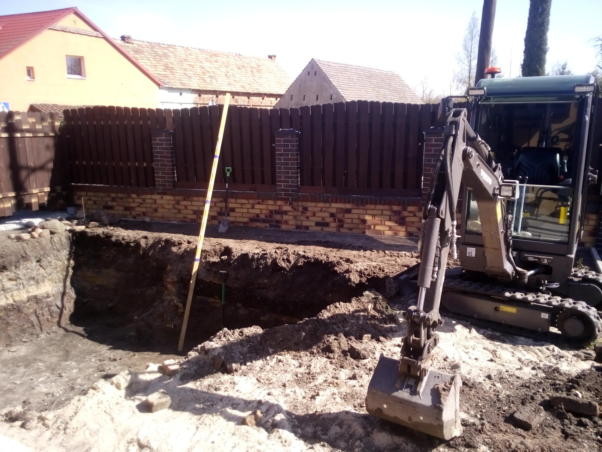 Mini koparka Volvo stoi obok wykopu w ziemi, widoczna poziomica i szpadel, w tle zabudowania gospodarcze i drewniany płot.