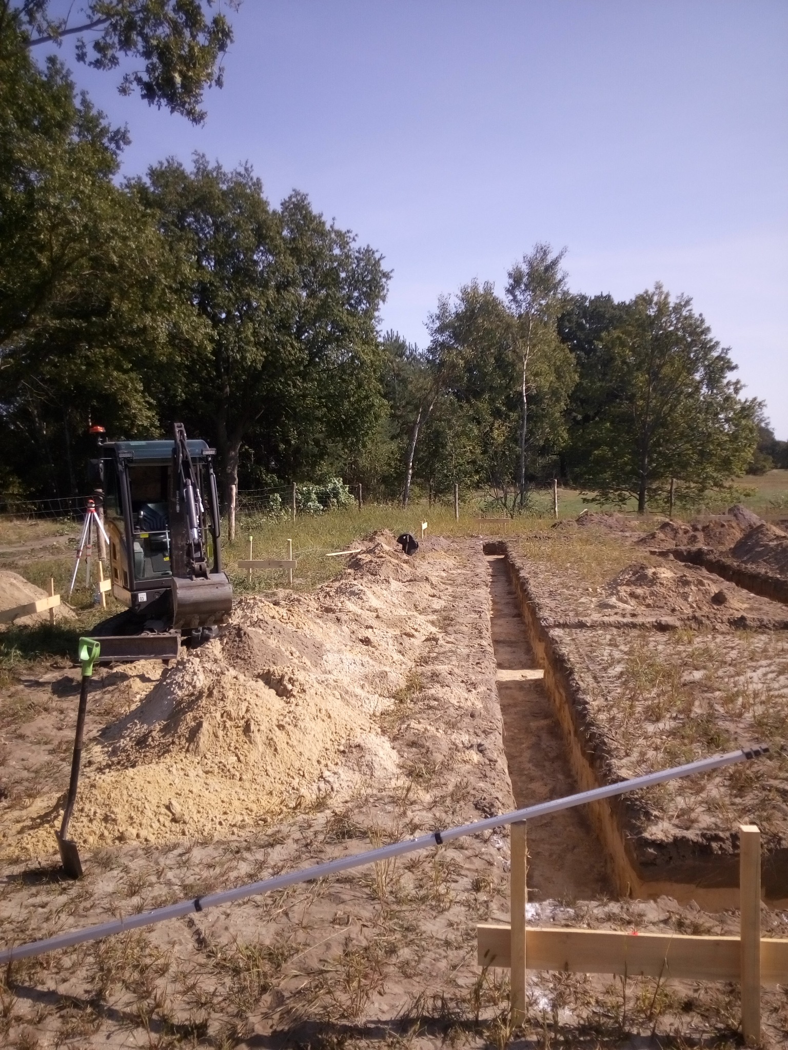 Mała koparka stoi obok wykopu pod fundamenty, widoczny niwelator laserowy i łata niwelacyjna, w tle drzewa i błękitne niebo.