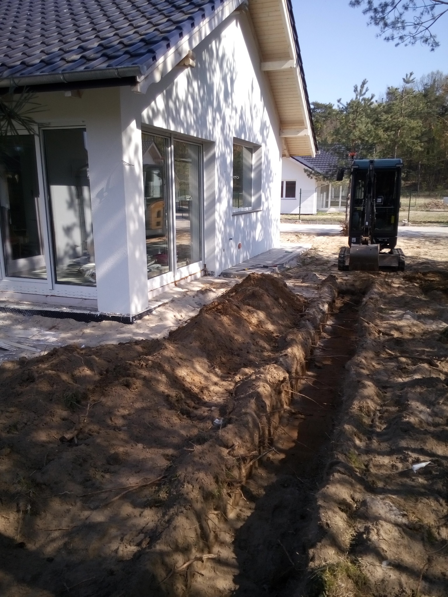 Wąski wykop wzdłuż fundamentów nowego, białego domu z dużymi oknami, z tyłu widoczny mały, ciemny traktor.