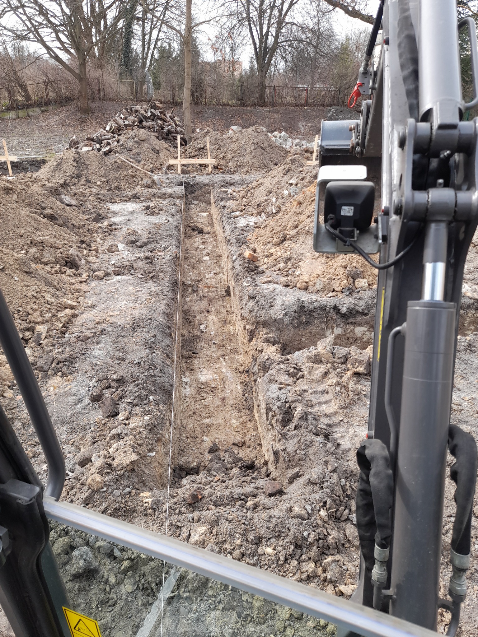 Wąski wykop w ziemi, widok z kabiny koparki, w tle pryzma ziemi i stos drewna.