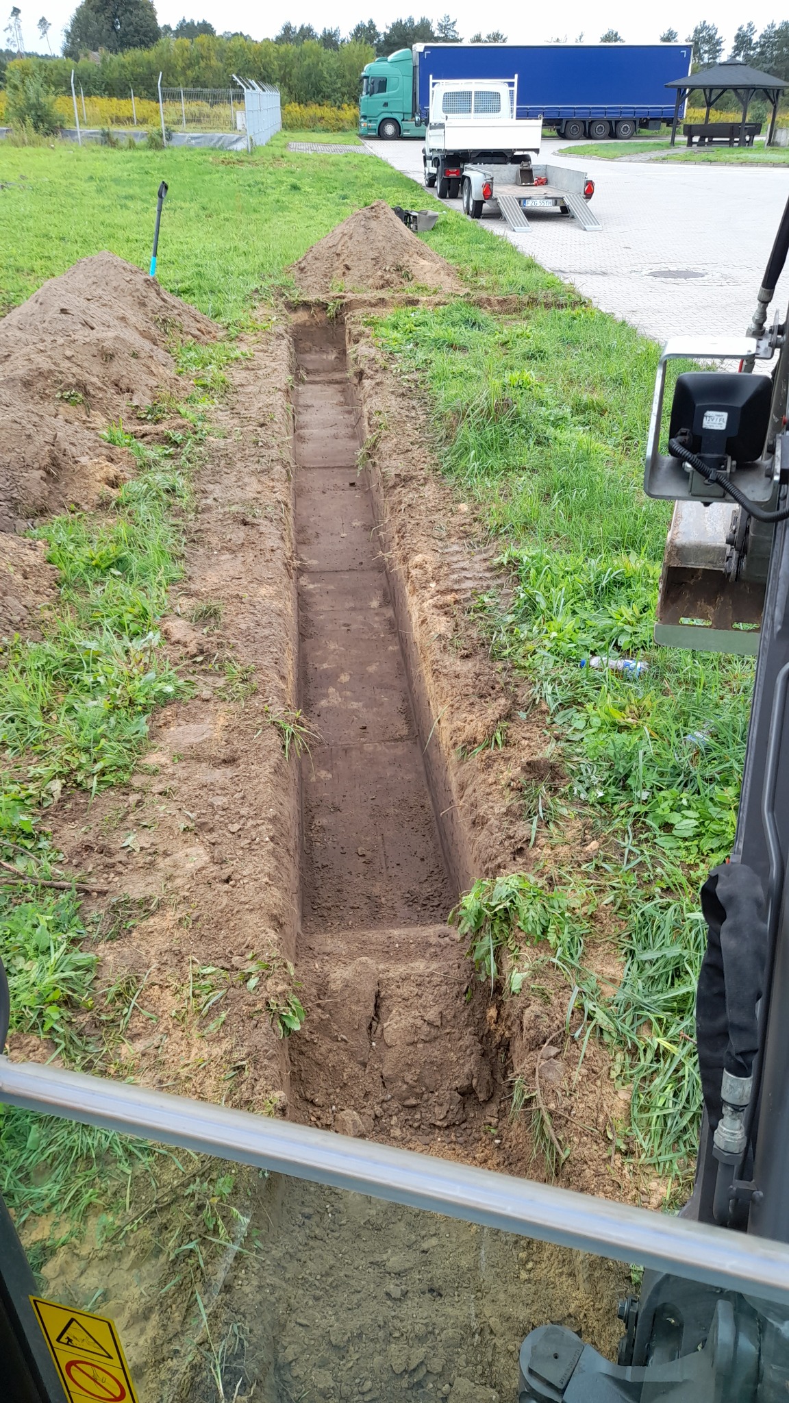 Wąski wykop liniowy w ziemi, widok z kabiny koparki, w tle zaparkowane ciężarówki i trawnik.