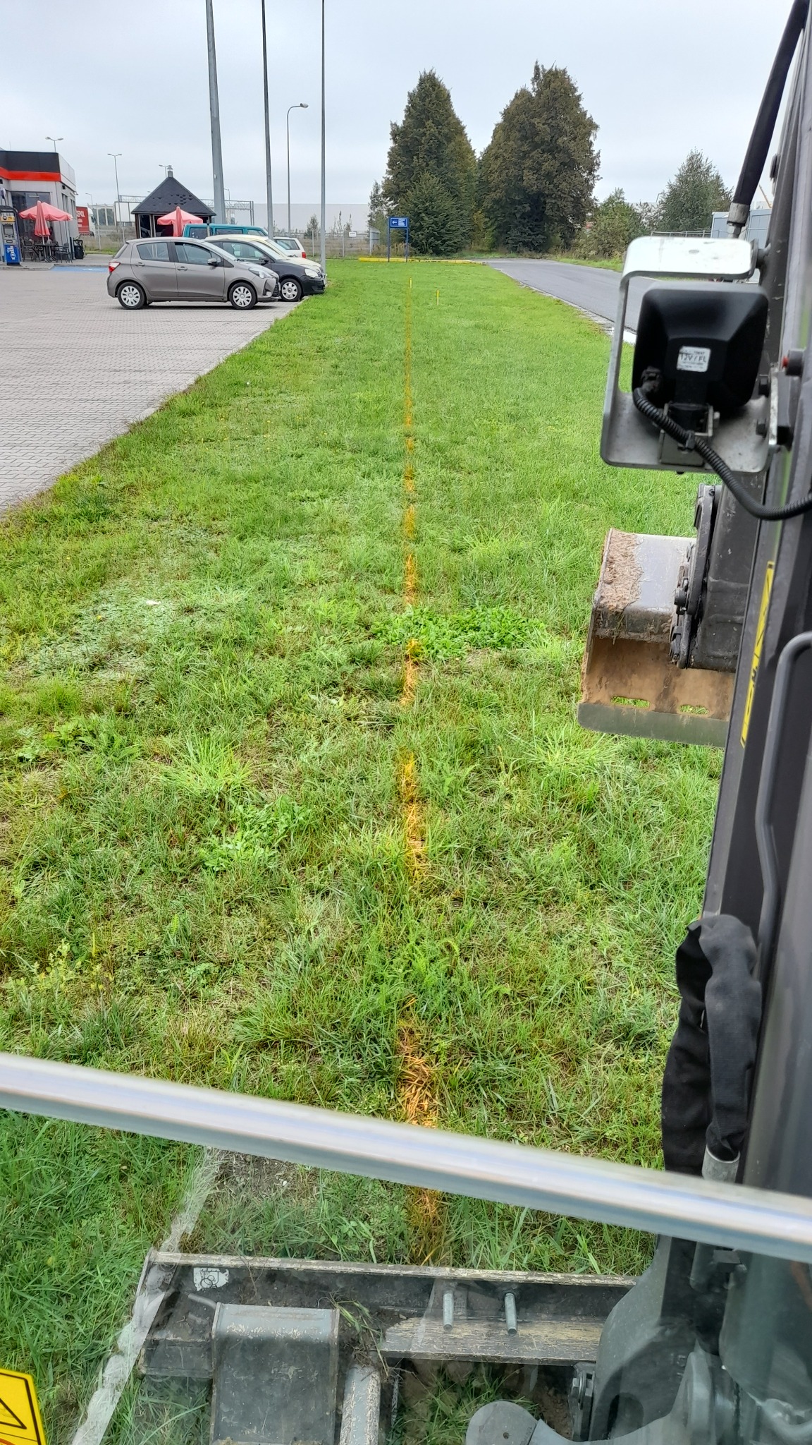 Żółta linia wyznaczająca trasę wykopu na trawniku, widok z kabiny koparki, w tle parking i drzewa.