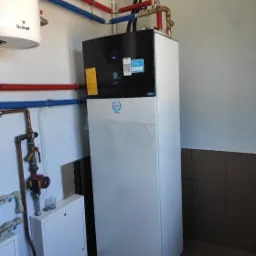 Biała pompa ciepła z czarnym panelem górnym, częściowo owinięta folią ochronną, z widocznymi rurami instalacji grzewczej w kolorach czerwonym i niebieskim oraz dodatkowymi elementami instalacji obok.