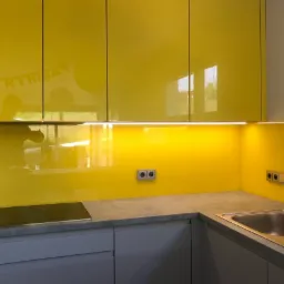 Żółty panel szklany w kuchni, podświetlony LED, z odblaskami, nad blatem z szarą powierzchnią, widoczny zlew i płyta indukcyjna.