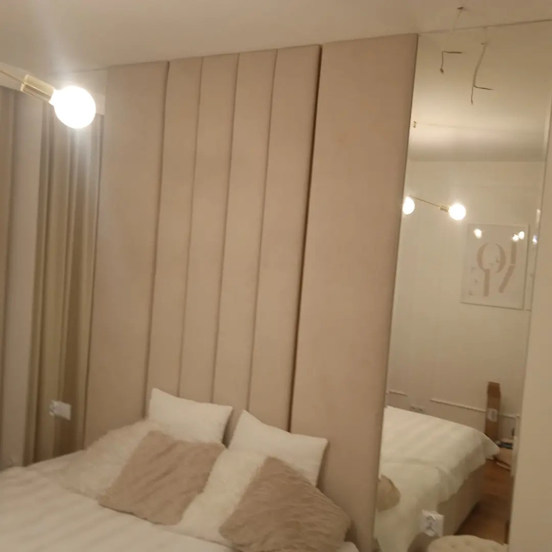 Beżowa tapicerowana ściana za łóżkiem z lustrem po prawej stronie, odbijającym część pokoju.