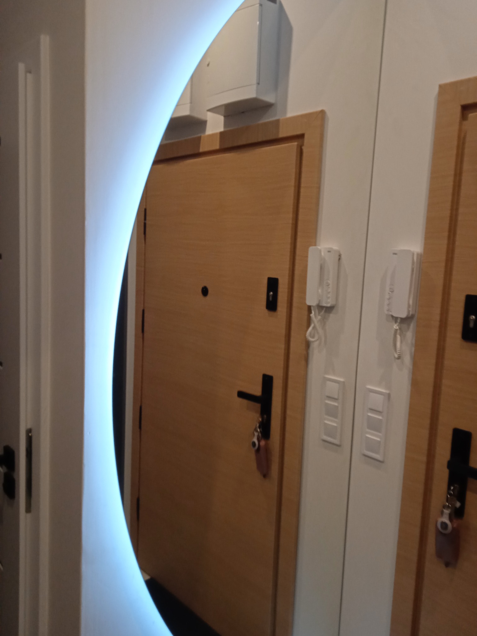 Lustro z podświetleniem LED odbija fragment dwóch drewnianych drzwi wejściowych z czarnymi klamkami i zamkami, obok domofonów na białej ścianie.