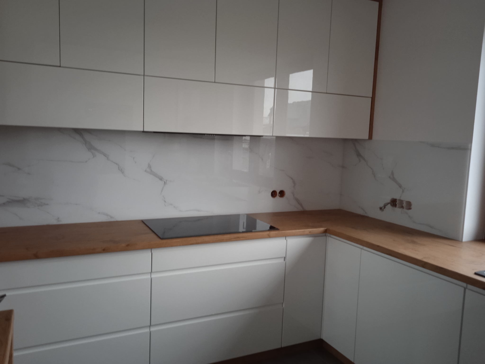 Nowoczesna kuchnia z białymi, lakierowanymi szafkami, drewnianym blatem i marmurowym panelem ściennym, widoczne przygotowane otwory na gniazdka elektryczne.