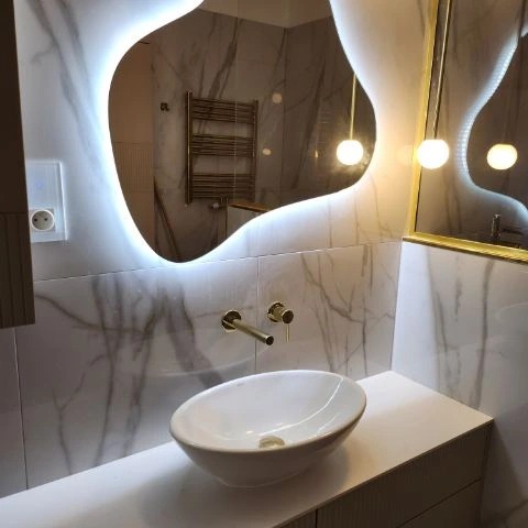 Nowoczesna łazienka z umywalką nablatową, baterią w kolorze szczotkowanego złota i asymetrycznym lustrem podświetlanym LED, odbijającym grzejnik drabinkowy.