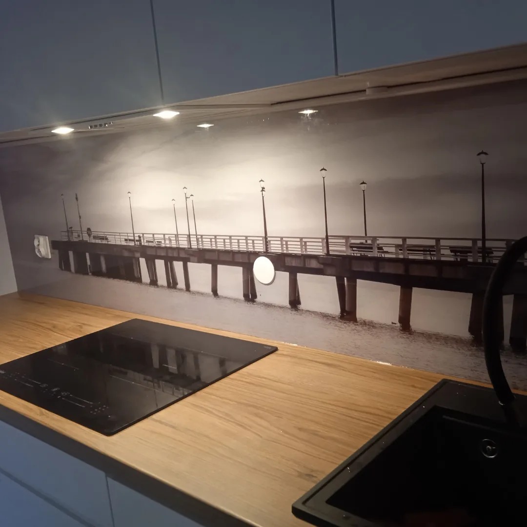 Szklana ściana nad blatem kuchennym z czarno-białym motywem molo, widoczna płyta indukcyjna i fragment zlewu.