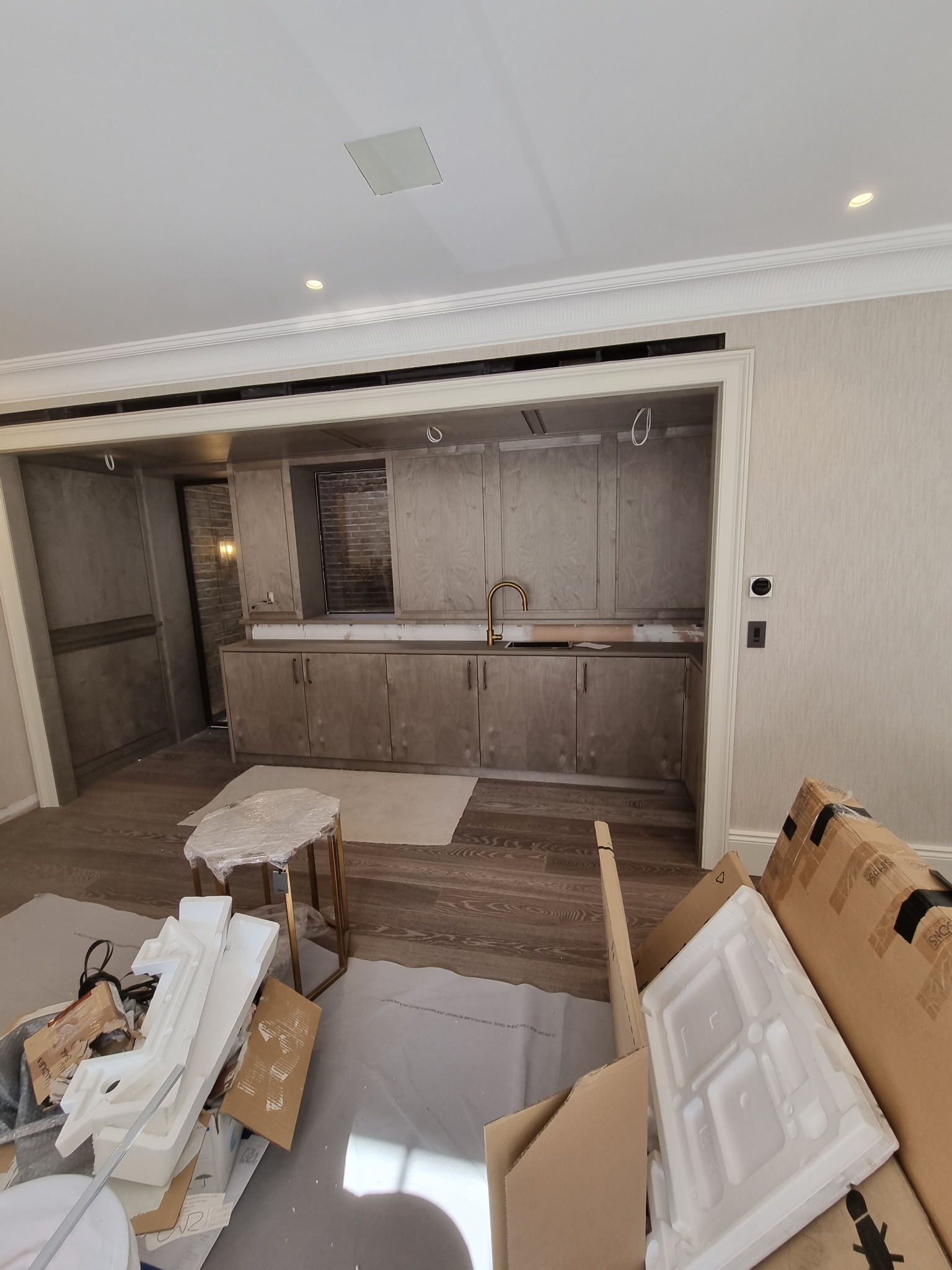 Wnętrze łazienki w trakcie prac wykończeniowych: zabudowa meblowa z szarymi frontami i złotą armaturą, widoczne kartony i zabezpieczenia podłogi.